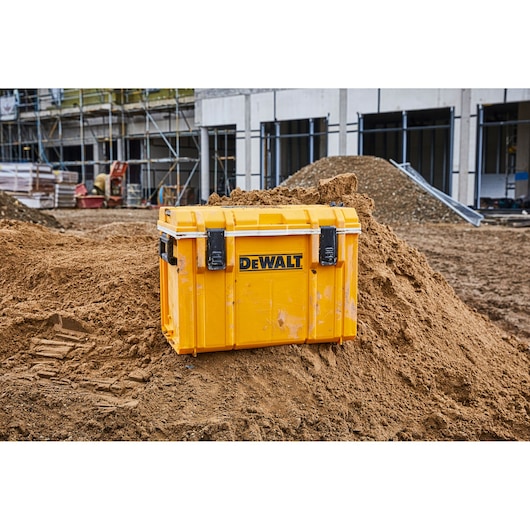 A yellow DEWALT tool storage box is placed on a mound of dirt at a construction site with scaffolding and unfinished buildings in the background.