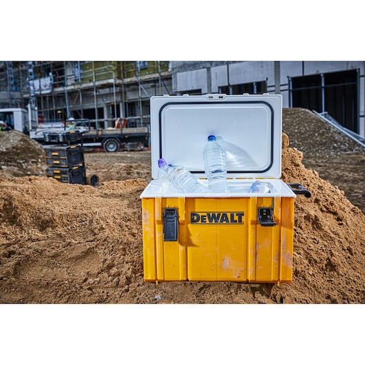 A yellow DEWALT cooler box with an open lid, containing plastic water bottles and ice, is placed on a construction site with dirt, construction materials, and buildings in the background.