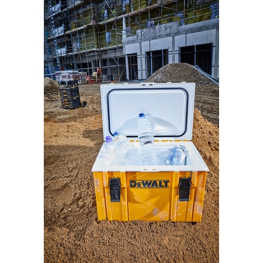 A yellow DEWALT cooler box with an open lid, containing several plastic water bottles and cans, placed on a construction site with scaffolding and building materials in the background.