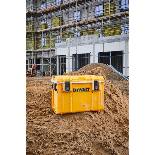 A yellow DEWALT toolbox is placed on a mound of dirt at a construction site, with a building and scaffolding visible in the background.