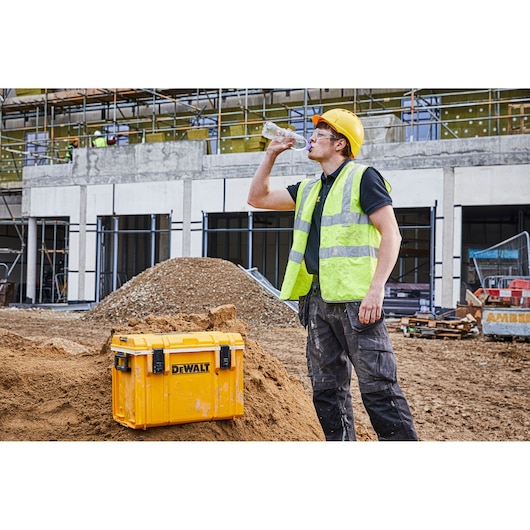 A construction worker wearing a yellow safety helmet and high-visibility vest drinks water from a plastic bottle next to a DEWALT tool case placed on a dirt mound at a construction site.