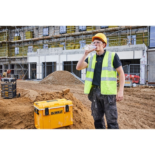A construction worker wearing a yellow hard hat and a high-visibility vest is drinking water at a construction site. The worker is standing next to a yellow DEWALT toolbox (DWST1-81333) placed on a pile of dirt. Scaffolding and a building under construction are visible in the background.