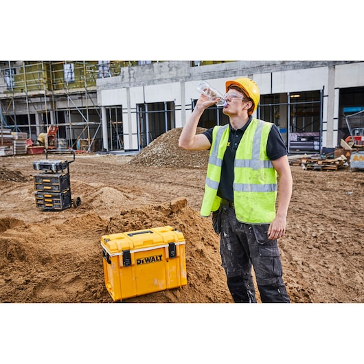 A construction worker wearing a yellow hard hat and high-visibility vest drinks from a plastic water bottle on a building site. A yellow DEWALT toolbox (SKU: DWST1-81333) is placed on a mound of dirt beside him. The environment features other toolboxes and construction materials with a building under construction in the background.