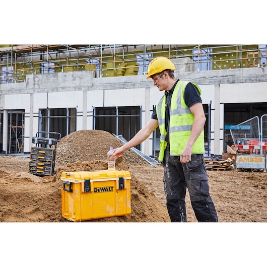 A blurred-face construction worker wearing a yellow hard hat and high-visibility vest stands at a construction site. He is placing a water bottle on a bright yellow DEWALT storage box situated on a mound of dirt. The background shows building frames, scaffolding, and stacked toolboxes.