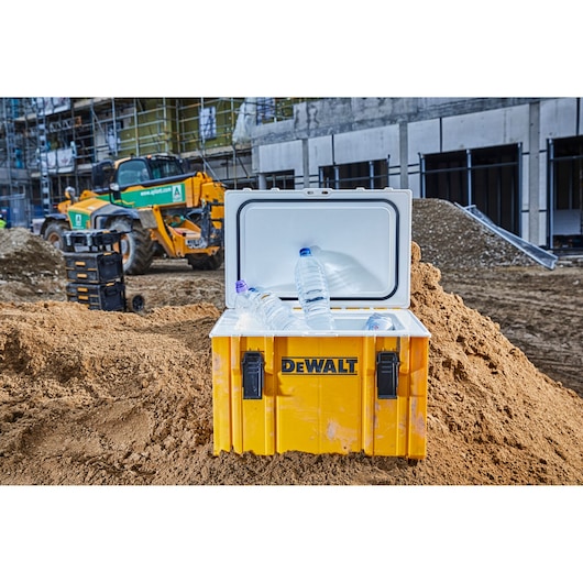 A yellow DEWALT storage box with an open lid containing several plastic water bottles, placed on a mound of dirt at a construction site. A construction vehicle and stacked storage boxes are visible in the background.