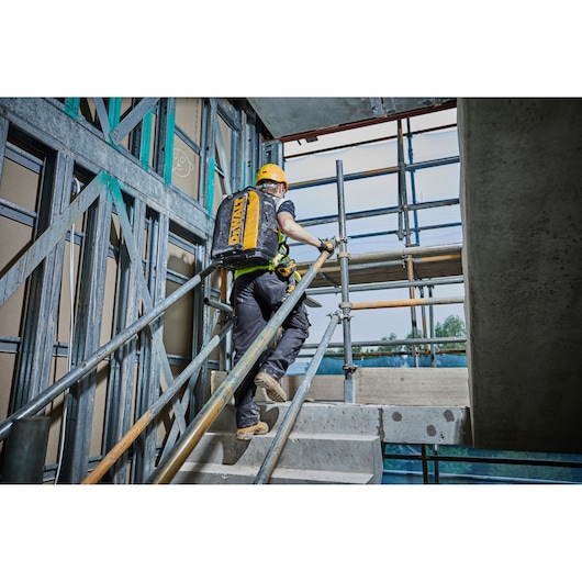 A person wearing a yellow hard hat and safety gear is ascending a concrete staircase at a construction site. The person is