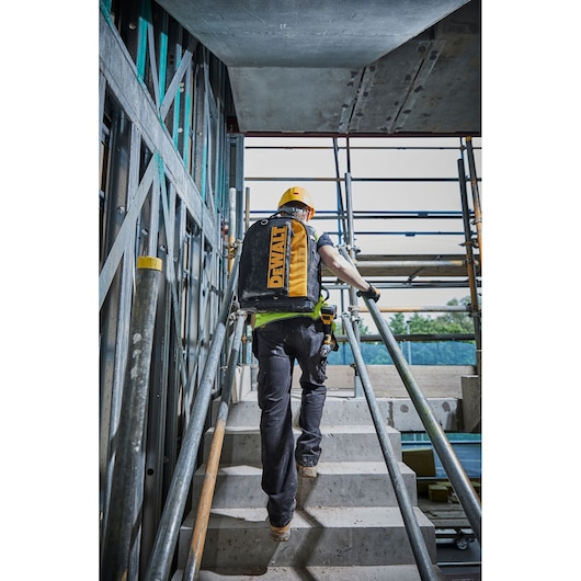 A person wearing a yellow hard hat and carrying a DEWALT branded backpack is walking up a concrete staircase at a