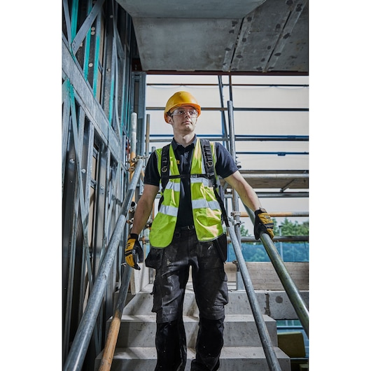 A construction worker wearing a yellow safety helmet, reflective vest, black work trousers, and gloves stands on a concrete