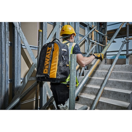 A person wearing a yellow hard hat, safety vest, and work gloves is carrying a DEWALT-branded backpack while ascending a