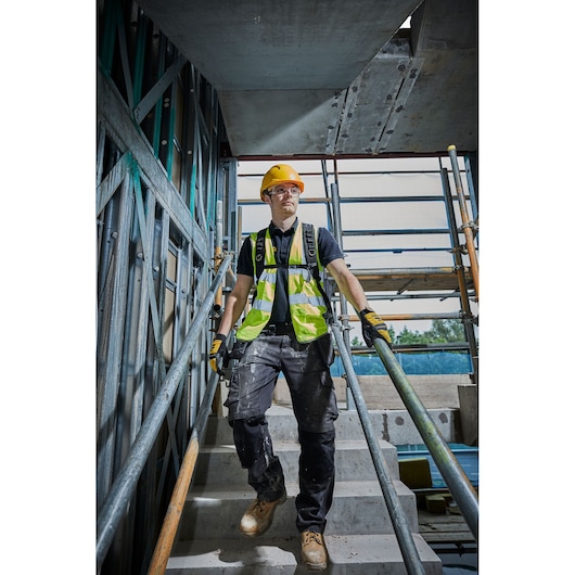 A construction worker wearing a yellow hard hat, high-visibility vest, gloves, and work boots is walking down a concrete