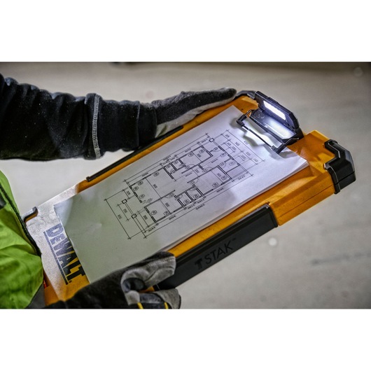 A person wearing gloves holds a DEWALT TSTAK clipboard with an integrated LED light, displaying a technical floor plan drawing attached to the clip.