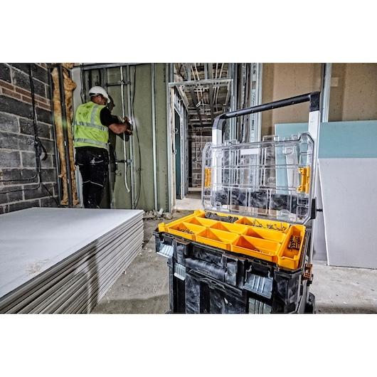 A DEWALT DWST82968-1 mobile tool storage box with open lid showing organized compartments filled with fasteners and hardware. The box is placed at a construction site, with drywall sheets stacked on the floor and a worker wearing a safety vest and helmet installing pipes in the background.
