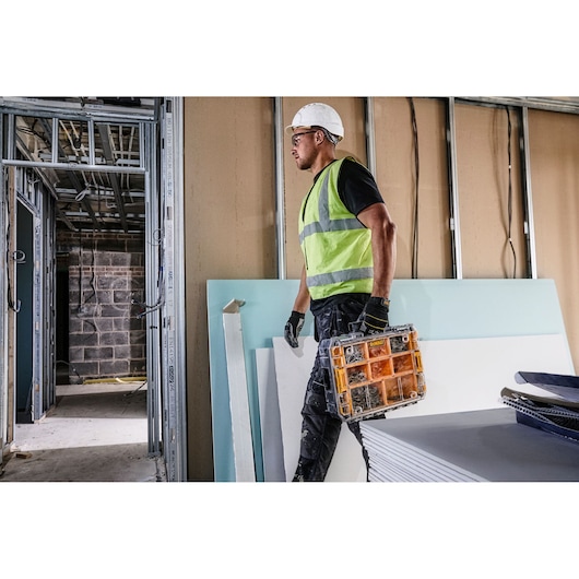 A construction worker wearing a high-visibility vest and white hard hat carries a DEWALT DWST82968-1 organizer case in a building under construction.