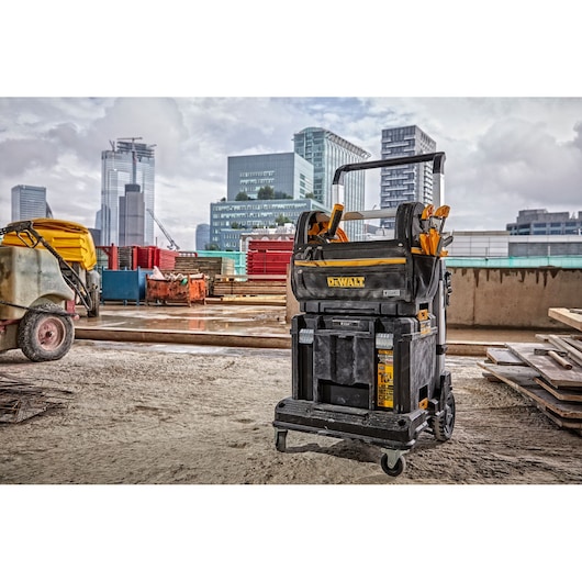 A DEWALT mobile tool storage unit (DWST82990-1) equipped with multiple compartments and hand tools, positioned on a construction site with modern city buildings in the background.