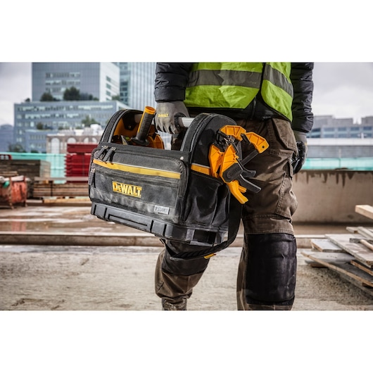 A person wearing work gloves and a green safety vest carries a Dewalt tool bag filled with various tools, including clamps, on a construction site with city buildings in the background.