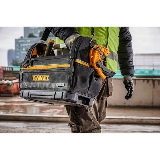 A person wearing work gloves and a high-vis jacket is carrying a DEWALT tool bag with the logo visible. The bag is black with yellow accents, filled with tools, and has sturdy compartments. The scene appears to be on a construction site.