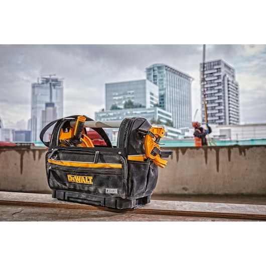 A DEWALT branded black and yellow tool bag with various tools is placed on a construction site surface, with modern city buildings and a construction worker blurred in the background.