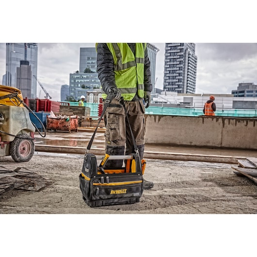 A DEWALT tool bag with multiple compartments is being carried by a worker wearing work gloves and a high-visibility vest on a construction site with urban buildings and equipment visible in the background.