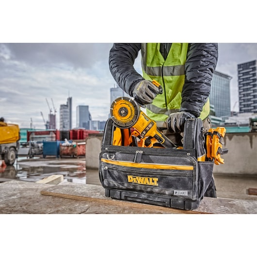 A person wearing work gloves and a high-visibility jacket is organizing DEWALT power tools inside a DEWALT TSTAK tool bag on a construction site, with tall city buildings and equipment in the background.