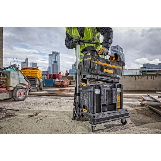 A person wearing a safety vest is handling a DEWALT tool bag and stackable rolling toolboxes at a construction site. Modern city buildings are visible in the background.