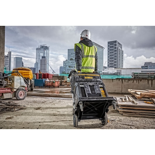 A person wearing a safety vest and hard hat is pulling a DEWALT DWST82990-1 rolling toolbox on a construction site with city buildings in the background.