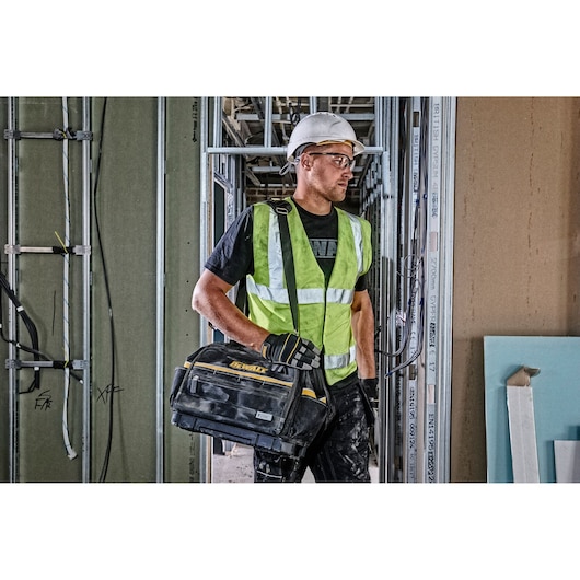A person wearing a reflective safety vest, hard hat, and gloves is carrying a DEWALT tool bag on a shoulder strap inside a