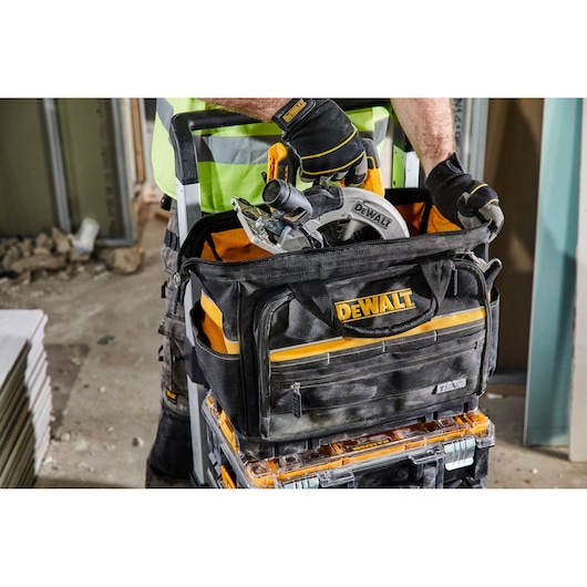 A close-up of a DEWALT branded tool bag, filled with tools including a circular saw. The bag is being carried by a person