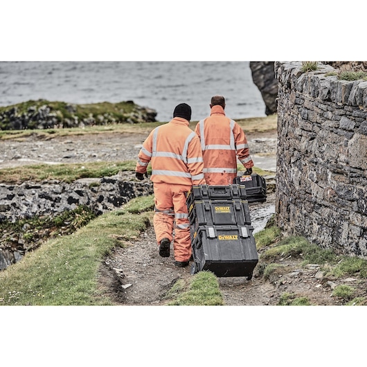 Two workers in orange high-visibility clothing are outdoors, walking along a dirt path near a stone wall. They are pulling