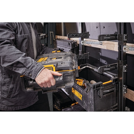 A person placing a DEWALT ToughSystem tool box onto a rack inside a workshop or vehicle storage area. Several DEWALT