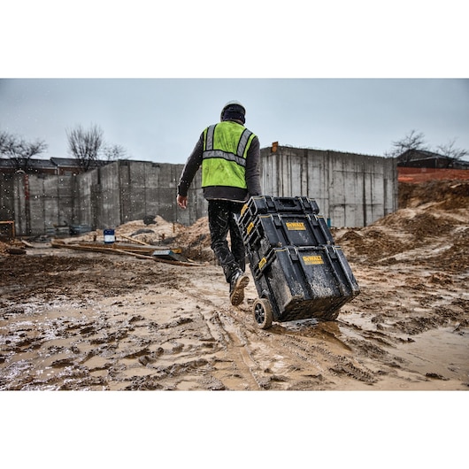 A person wearing a green safety vest and helmet is pulling a DEWALT rolling toolbox through a muddy construction site, with