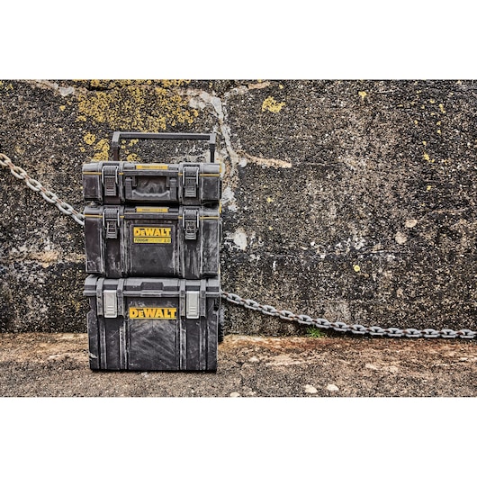 Three stacked DEWALT ToughSystem 2.0 toolboxes, black with yellow DEWALT logo, set outdoors against a rough stone wall and a