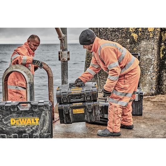 Two workers in orange high-visibility suits are outdoors by the waterfront, handling black DEWALT tool storage boxes. The