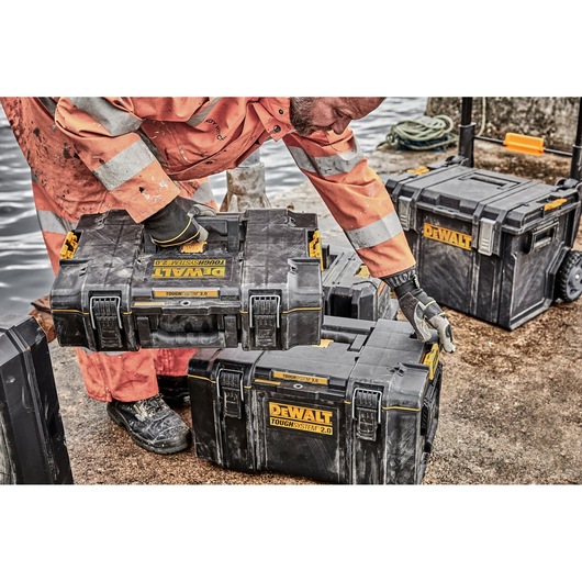 A person in high-visibility work clothing and gloves is handling a stack of DEWALT ToughSystem 2.0 toolboxes at an outdoor