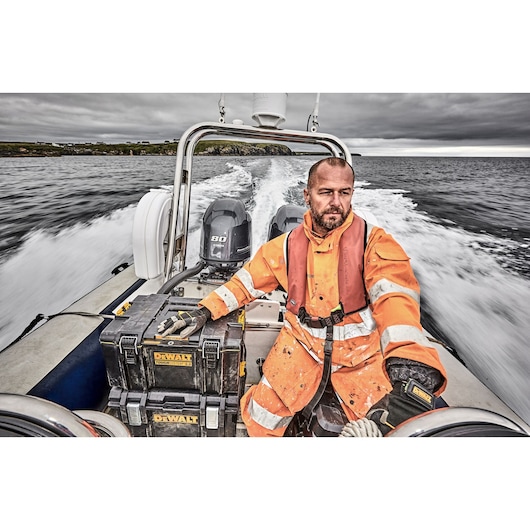 A person wearing an orange high-visibility waterproof suit and gloves is sitting on a motorboat. There are two black DEWALT