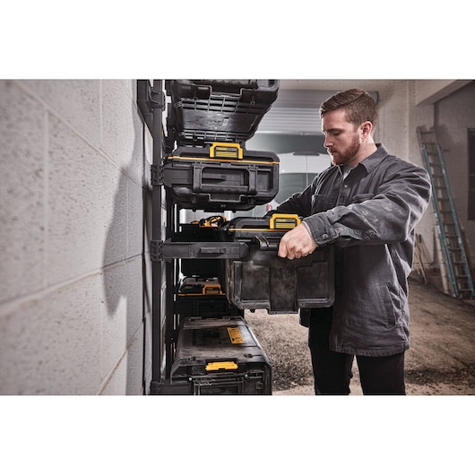 A person is organizing black and yellow DEWALT toolboxes on a wall-mounted shelving system in a workshop. The person's face