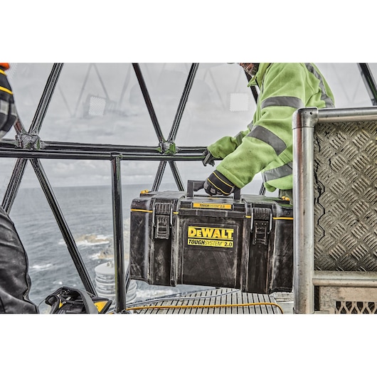 A person wearing a high-visibility jacket is holding a DEWALT ToughSystem 2.0 tool box inside a metal structure with a view