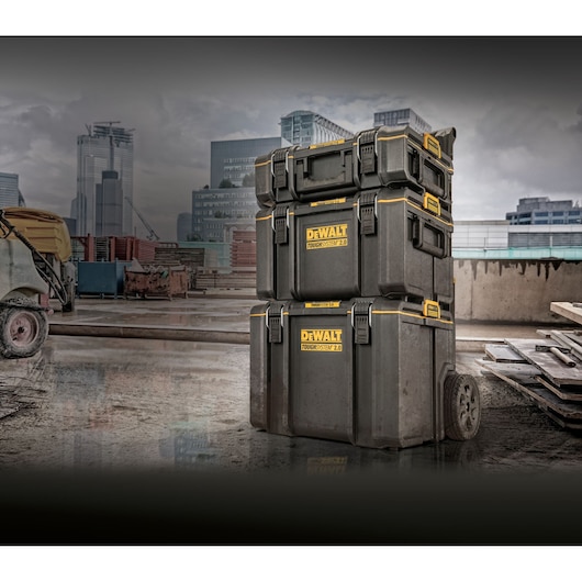 Three stacked DEWALT ToughSystem 2.0 tool storage boxes are shown on a construction site. The boxes are black with yellow accents and wheels for mobility. Modern city buildings and construction equipment are visible in the background.
