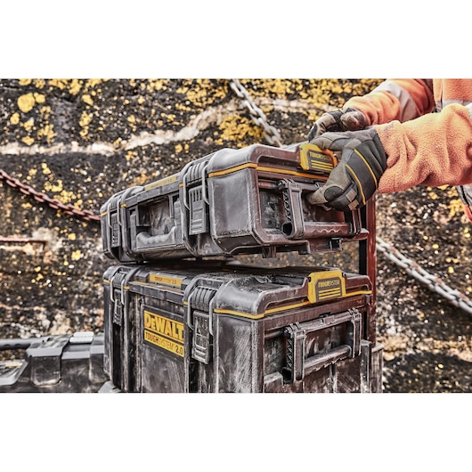 Gloved hands stacking DEWALT ToughSystem 2.0 toolboxes outdoors against a wall with yellow marks.