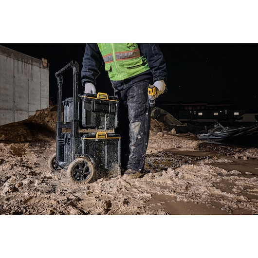 A person wearing a green safety vest and gloves stands on a muddy construction site at night beside stacked DEWALT tool