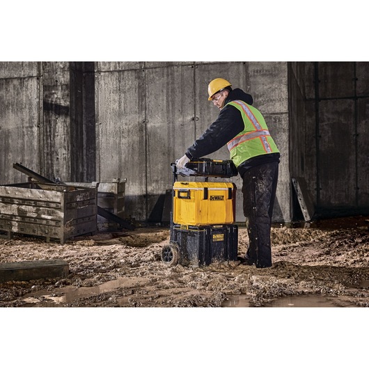 A person wearing a safety vest and hard hat is pushing a stack of DEWALT toolboxes with wheels across a muddy construction