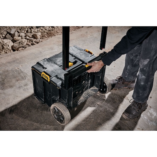 A person pulls a DEWALT rolling tool box with large rugged wheels and a telescopic handle on a dusty construction site.