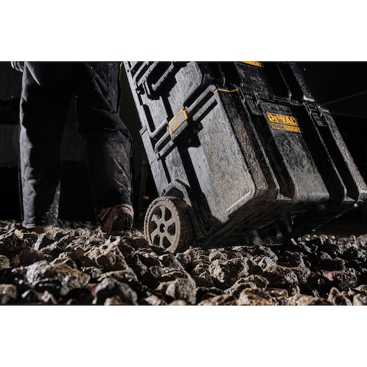 Close-up view of a person pulling a muddy DEWALT ToughSystem 2.0 wheeled tool box over rough rocky ground.
