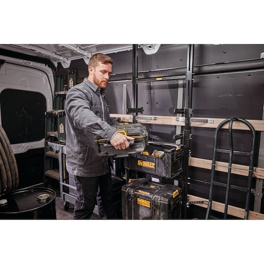 A person in a work jacket is organizing DEWALT toolboxes inside a van equipped with shelving and various equipment. The