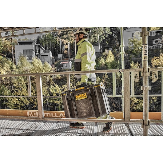 A person wearing high-visibility workwear and a safety helmet is carrying a DEWALT ToughSystem 2.0 storage box on a
