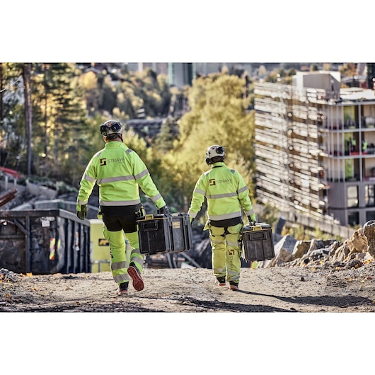 Two construction workers wearing high-visibility jackets and helmets are walking on a dirt path at a construction site, each