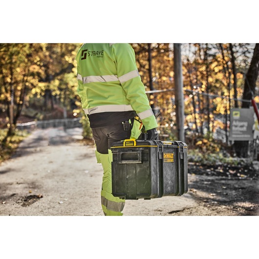 A person wearing high-visibility work clothing carries a DEWALT DWST83342-1 black and yellow toolbox down a path outdoors