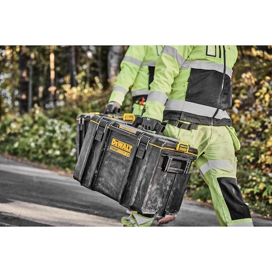 Two people in high-visibility work clothing carry a large black DEWALT ToughSystem 2.0 toolbox outdoors.