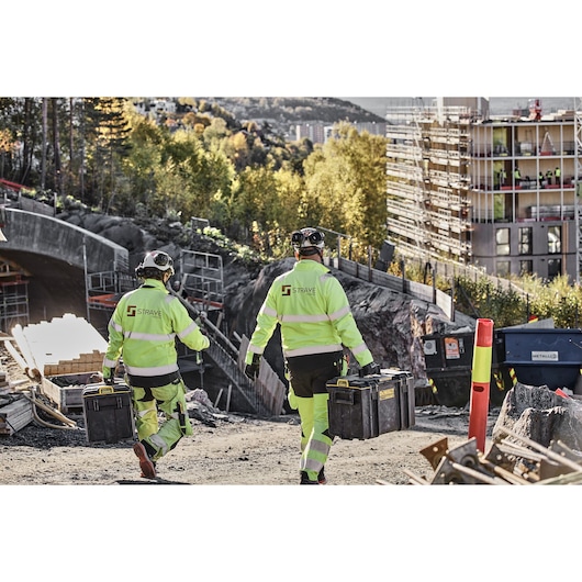 Two construction workers wearing high-visibility jackets and helmets walk at a construction site, each carrying a DEWALT