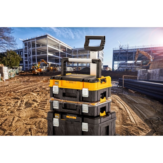 Stacked DEWALT TSTAK storage boxes with a telescopic handle, positioned on a construction site with buildings and machinery