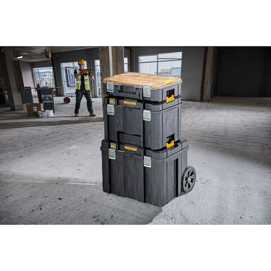A stack of three DEWALT black and yellow wheeled toolboxes is shown on a construction site floor, with a worker in a safety